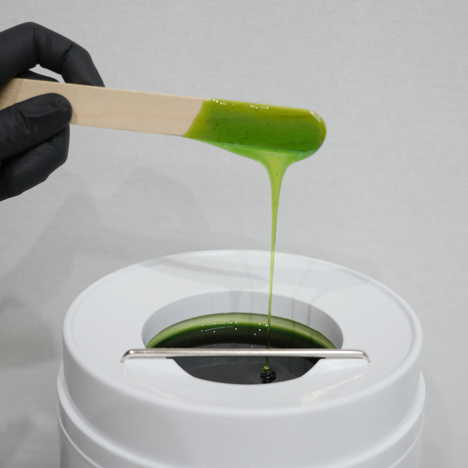 A hand wearing a black glove holds a wooden spatula above a wax pot, showing Caronlabs Smooth & Remove strip wax being dripped into the wax heater.