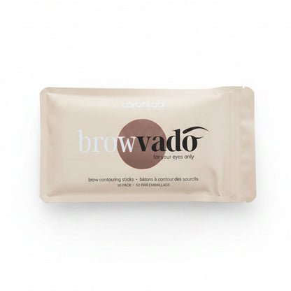 A single brow contouring stick packet by caronlab browvado, in a light beige foil wrapper with a brown oval logo, placed on a dark black background.