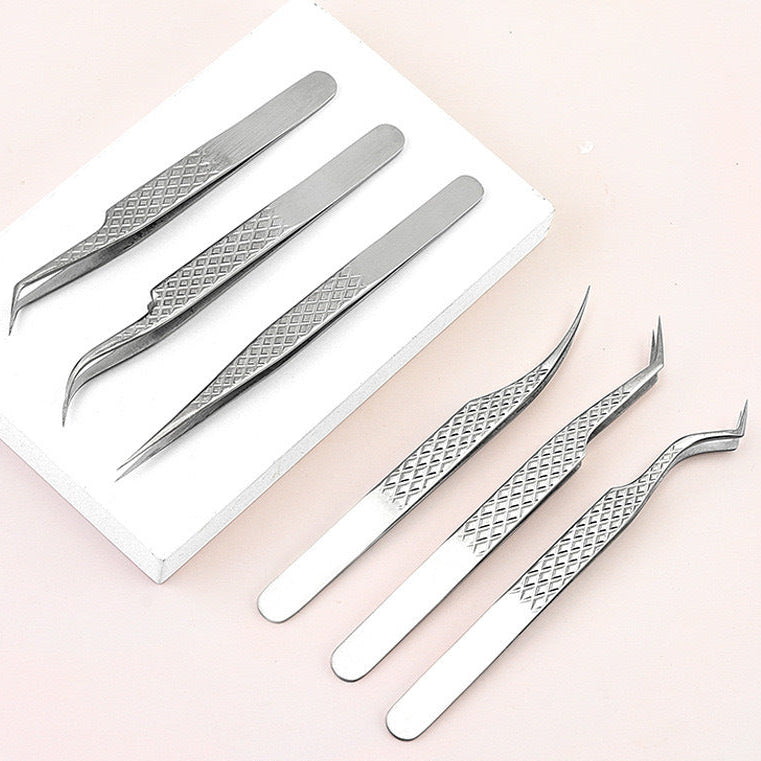 Five pairs of stainless steel precision tweezers with textured diamond-patterned handles, arranged on a white rectangular block against a soft pink background.